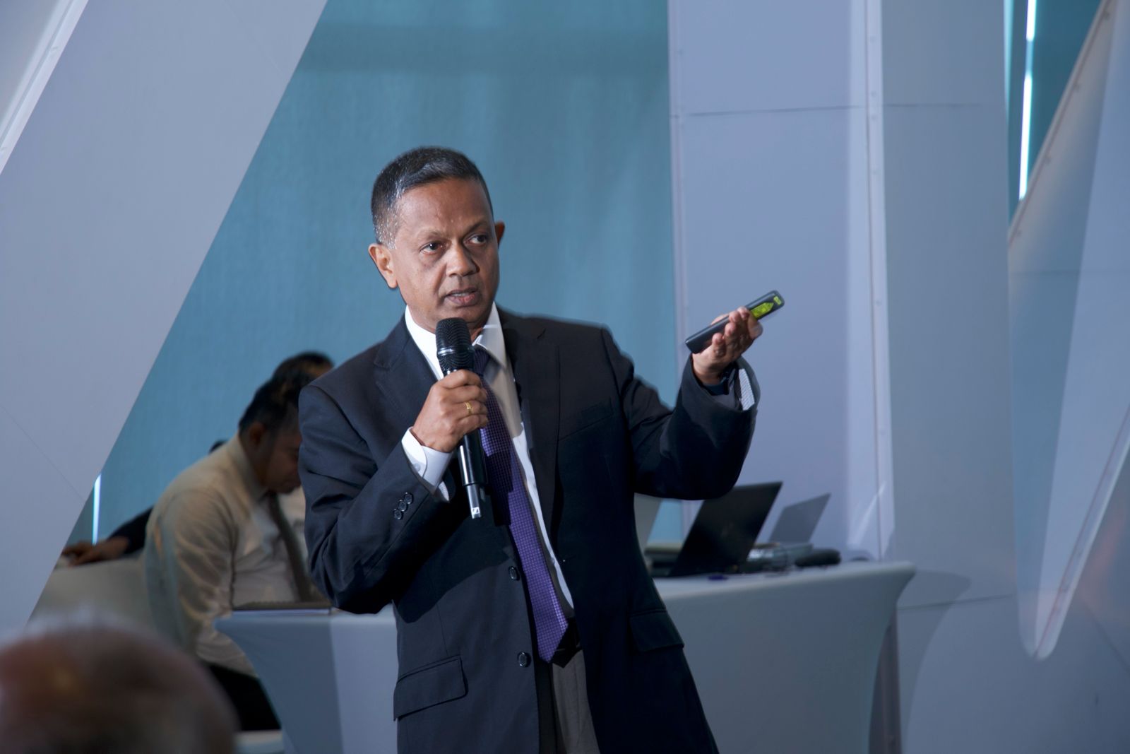 Sumith Jayawickrama speaking on stage — dark suit and tie, microphone in one hand, presenter remote in the other.
